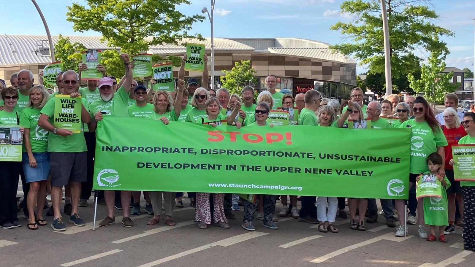 A large group of people standing holding a green banner. They are outside and are mostly wearing green T-shirts.
