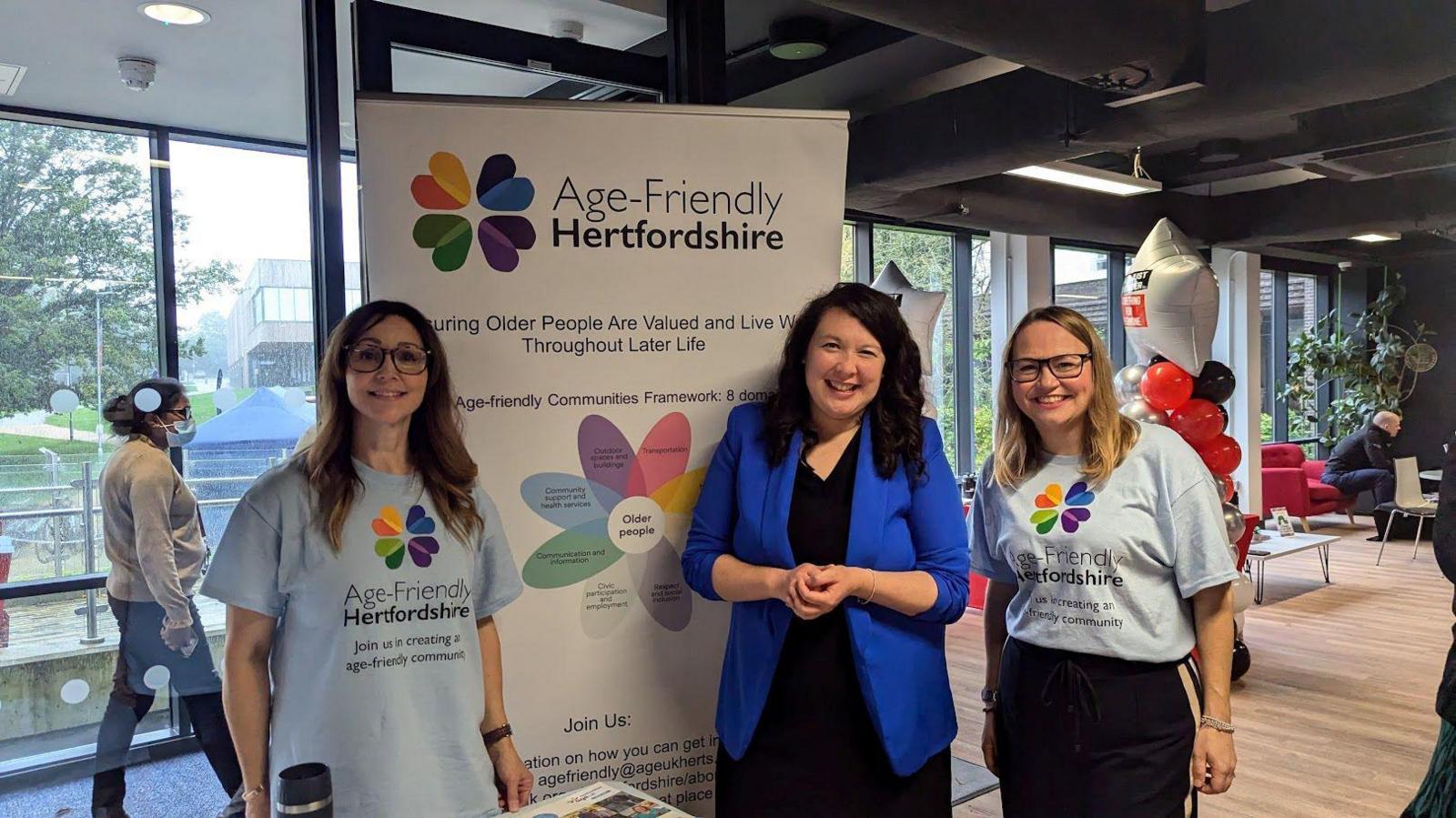 MP wearing a royal blue jacket and black dress is with two women from charity Age-Friendly Hertfordshire. They are stood wither side of the MP with pale blue t-shirts with the charity's logo on the front. There are people milling in the background at what looks like an opening event.