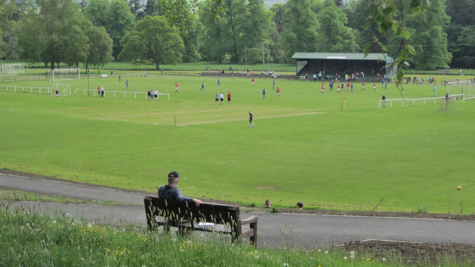A man on a bench looks down over a football field in the distance with a distinctive green stand