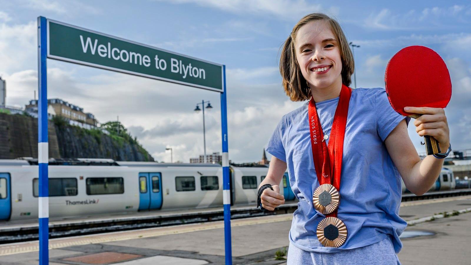 Bly Twomey holds a table tennis racket and wears her medals standing in front of a sign saying "Welcome to Bly-ton"