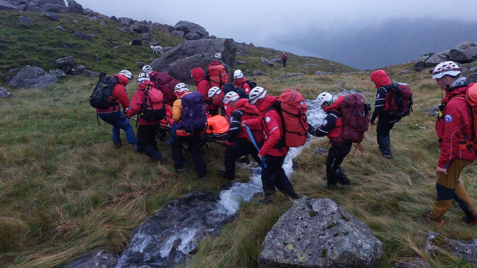 Wasdale Mountain Rescue Team carrying the casualty on a stretcher. A group of several people are wearing red jackets and white helmets. The are crossing a small stream on a hillside.