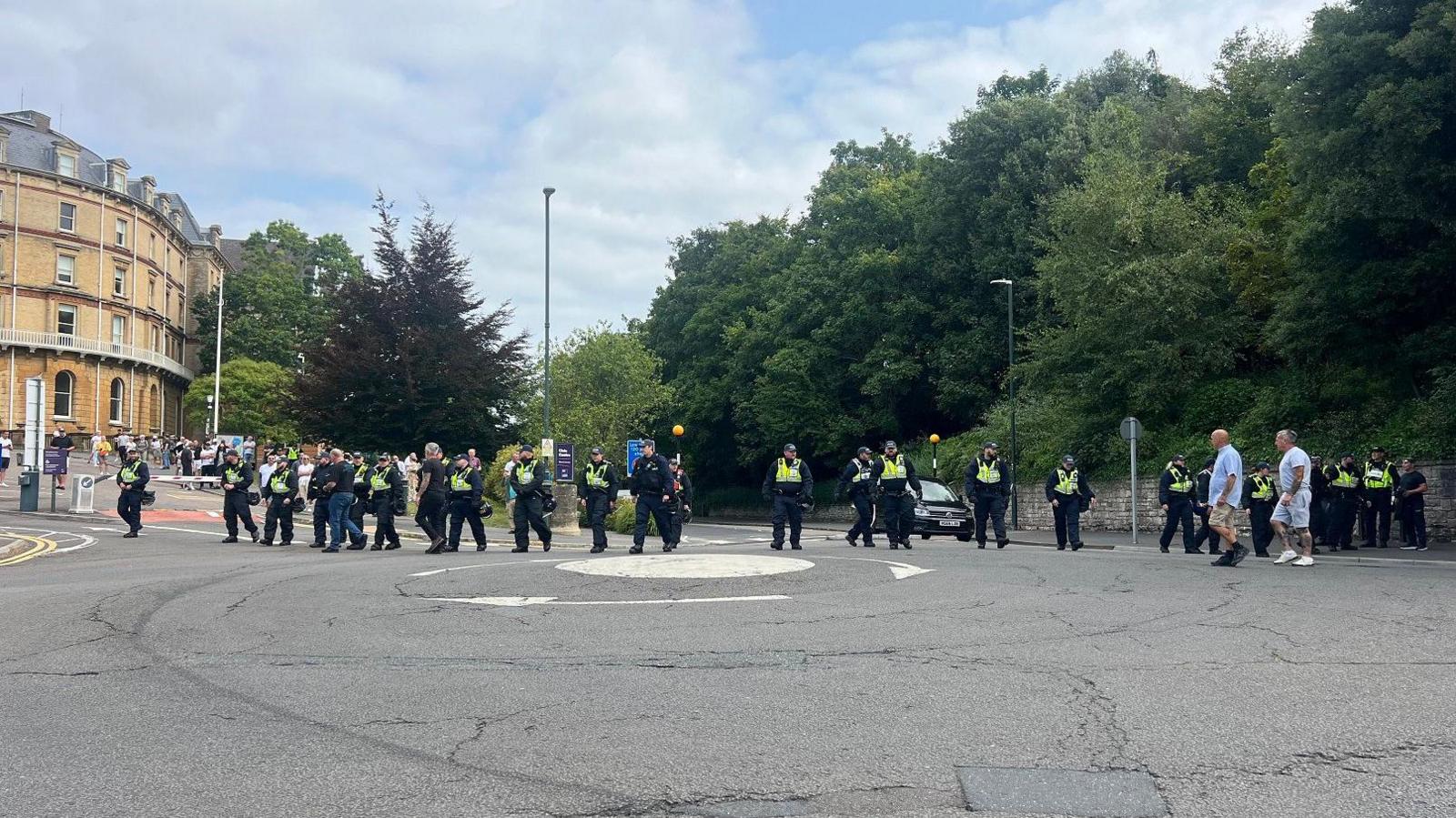 About 20 police officers standing at the edge of the road and walking across a roundabout. On the left hand side, the town hall building is just about visible.