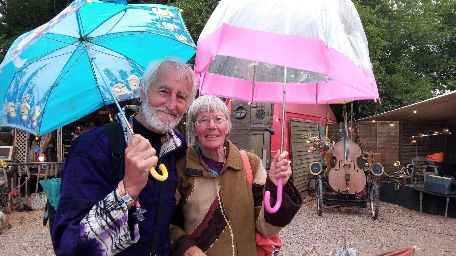 Graham and Judy Cole standing at Glastonbury Festival holding a blue and pink umbrella