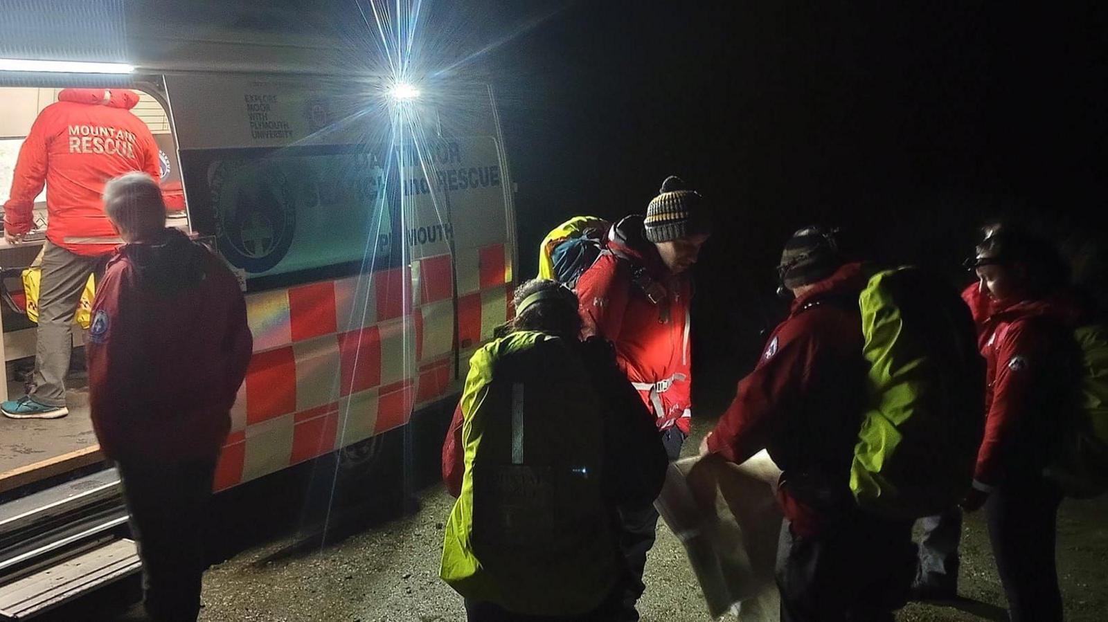 Members of Dartmoor Search and Rescue Team Plymouth gather outside of the rescue van on Tuesday evening. The team are wearing warm hats and coats. It is dark. One team member is inside the van looking at a computer screen which appears to show a map. Other members of the team are gathered around a paper map. A head torch is being used to illuminate the map.