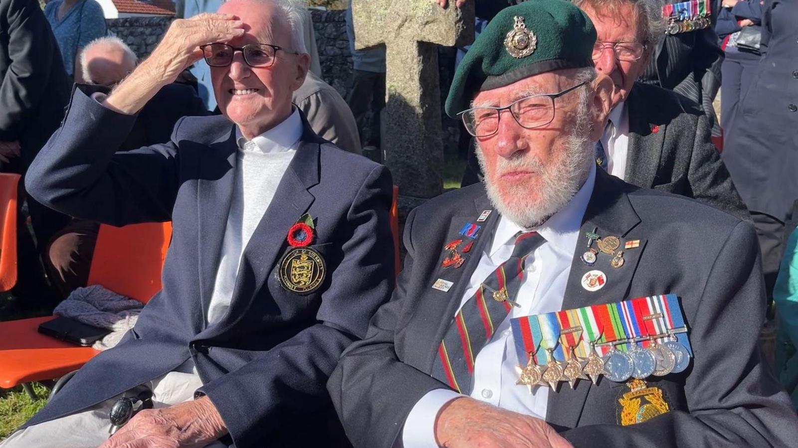 An old man in a blazer and green Royal Marines beret, with medals over his lapels. He sits on a chair next to other people, many in similar outfits.