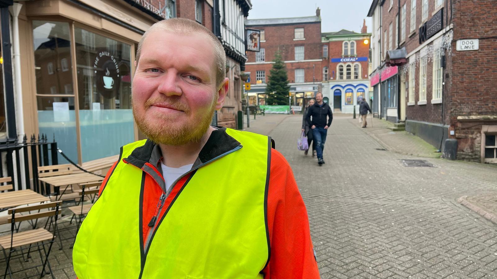 A man with cropped fair hair and stubble stands on a quiet tiled street with a few pedestrians in the background. He is wearing an orange coat covered by a yellow hi-vis vest. A cafe and outside tables are to the left of the image. A pub, shop fronts and a Christmas tree can be seen in the distance.