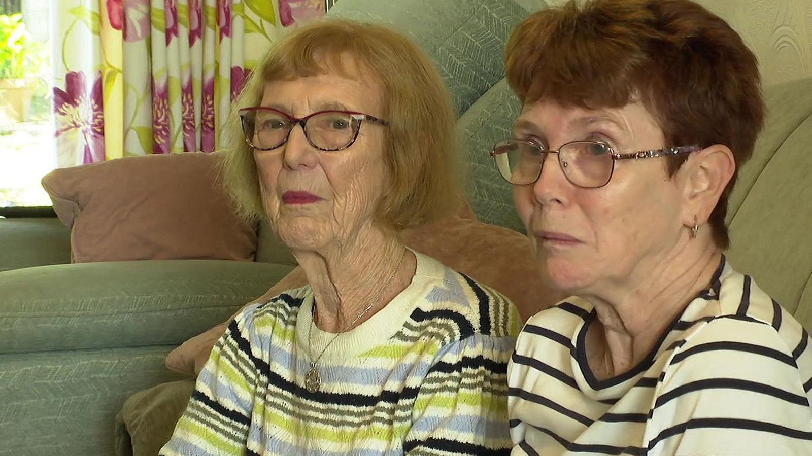 Mother and daughter Anne (left) and Louise Dixon sitting on a green sofa with flowery curtains hanging from the window behind them. Anne wears purple framed glasses and dark red lipstick with light brown bobbed hair and a white, green and grey striped jumper. Louise also wears glasses and has short, brown hair and a white-and-black striped top.