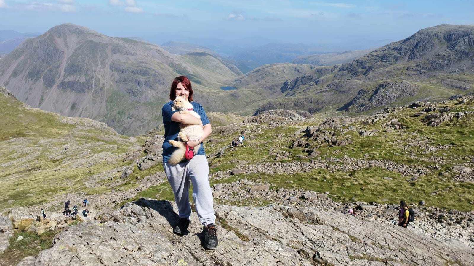 A woman in sports clothes and shoes holding a cat on a mountain path. It's a sunny day. A mountain panorama stretches behind them.