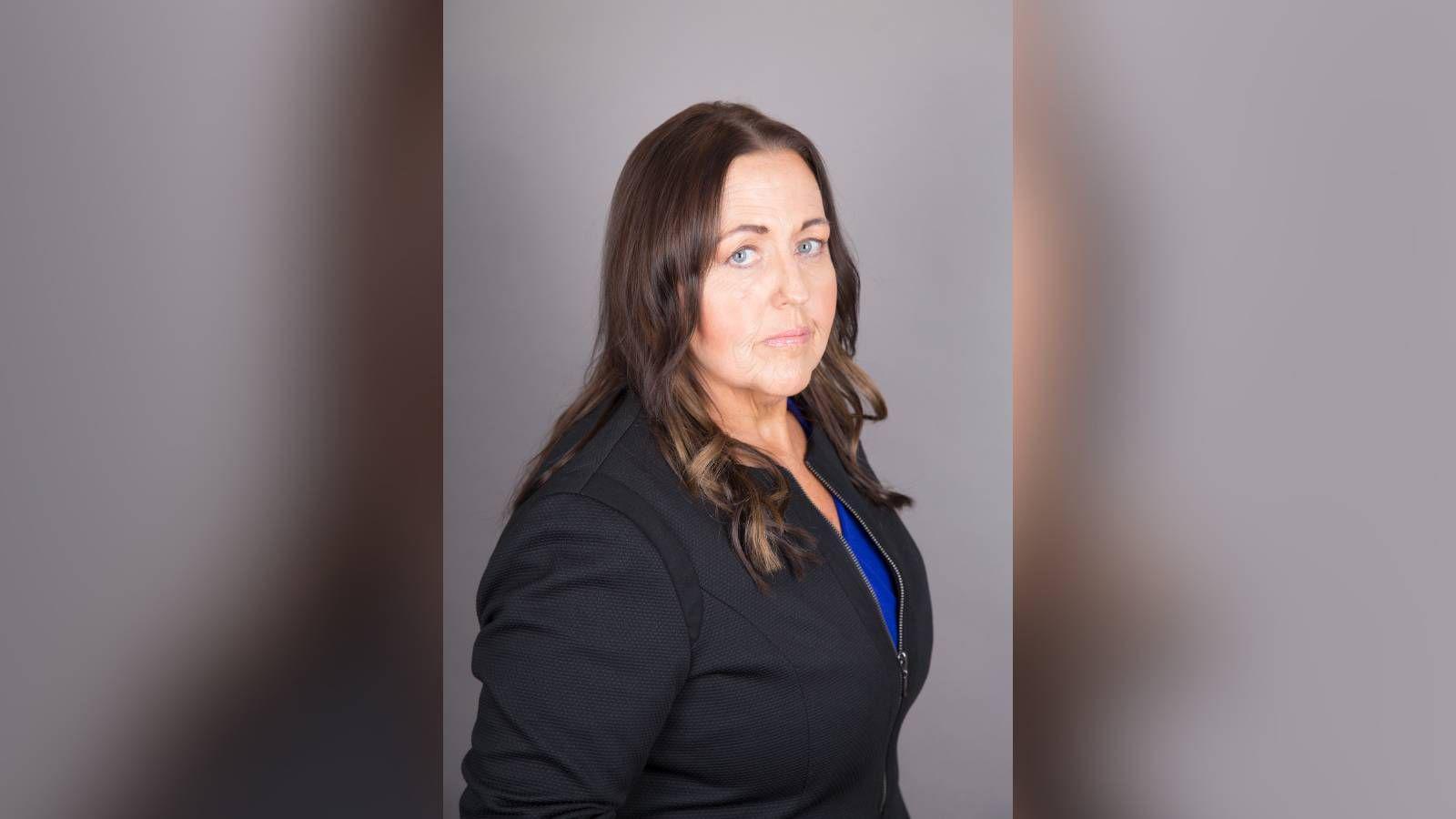 Pictured is Joanne Monk standing to the side with her head facing the camera. She has shoulder length brown hair