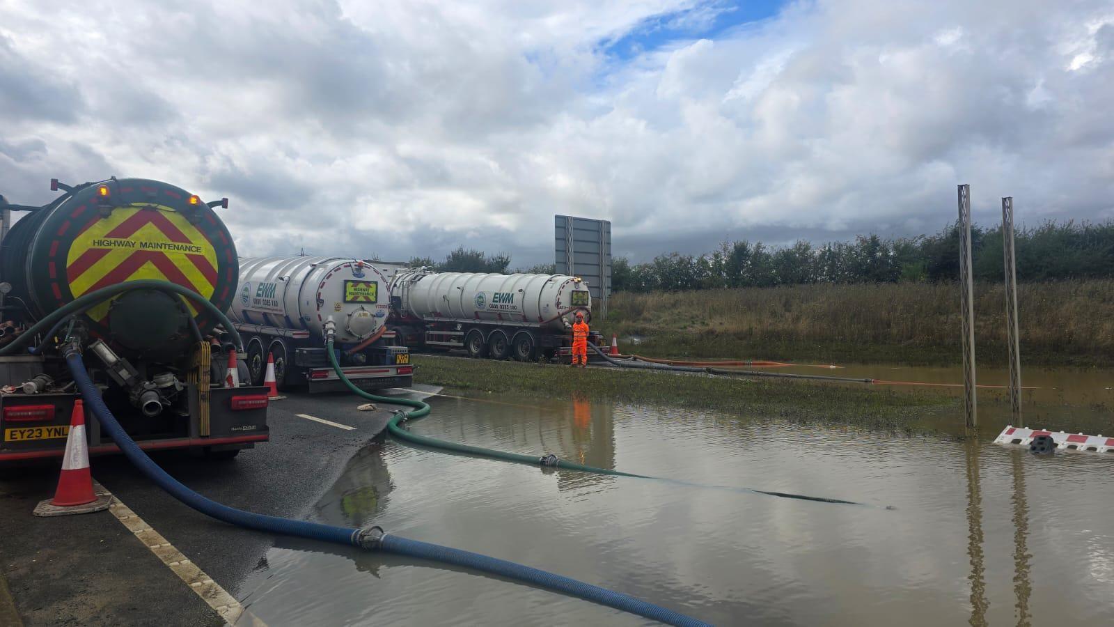 Three tankers on the A421, clearing water that is on the road surface, with a man standing in an orange florescent suit, by one