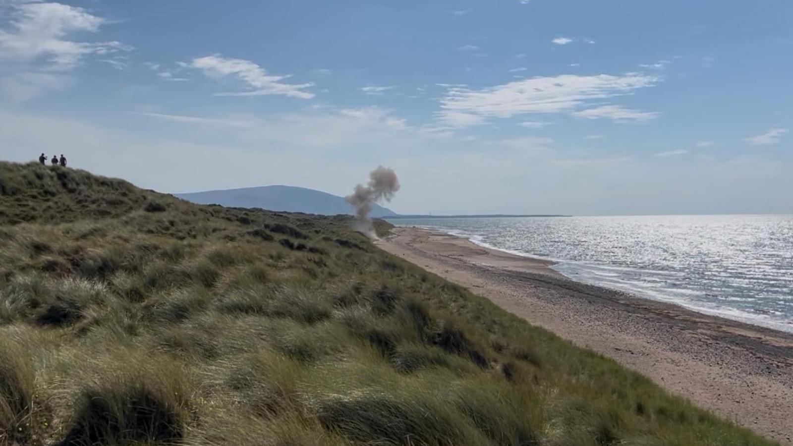 Long shot of smoke coming from beach on a sunny day. The photo is taken from a grassy field next to the beach and the ocean. The sun is reflecting off the ocean.