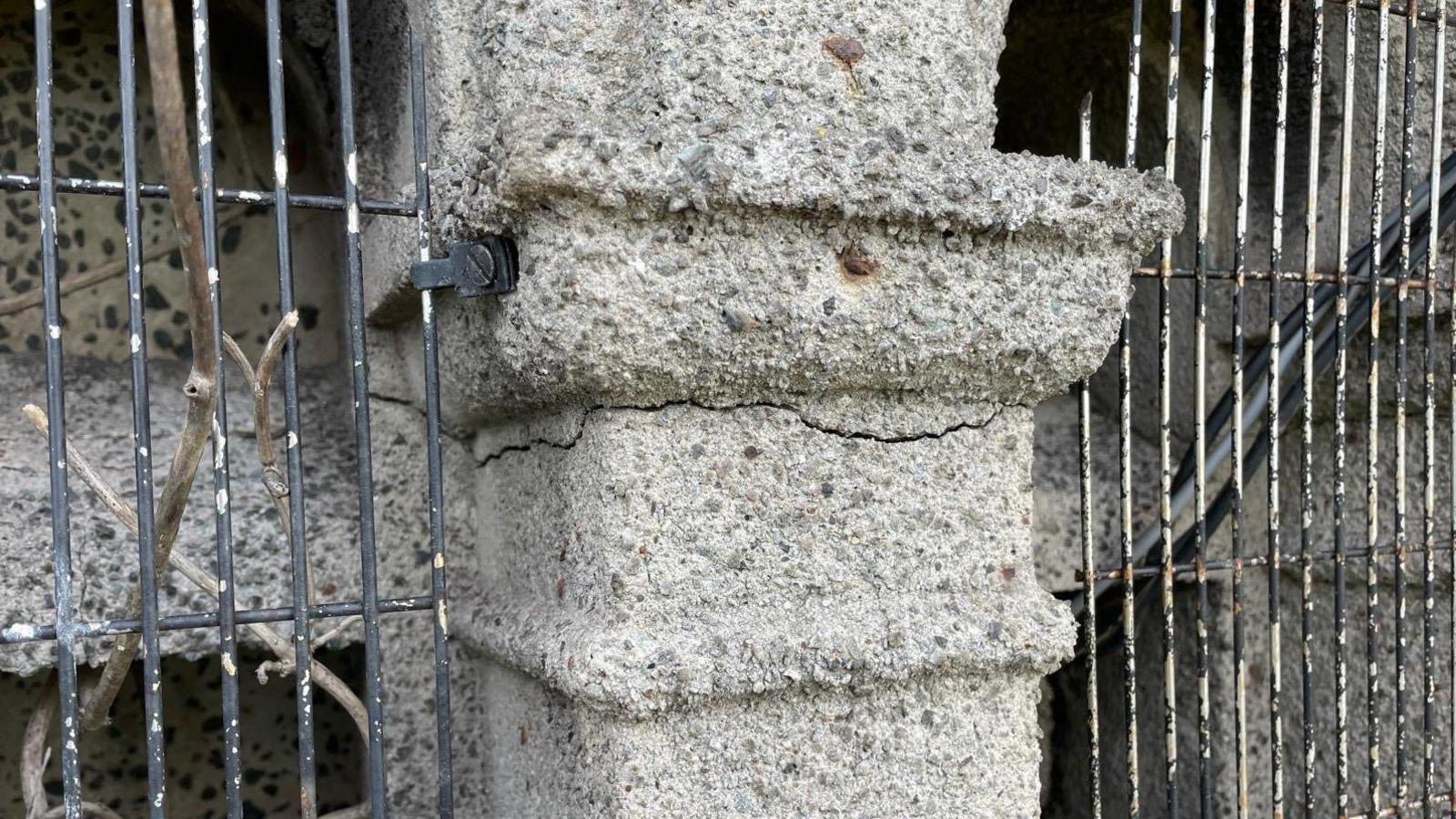 A close-up of a concrete pillar between two metal gates has a long horizontal crack running through it.