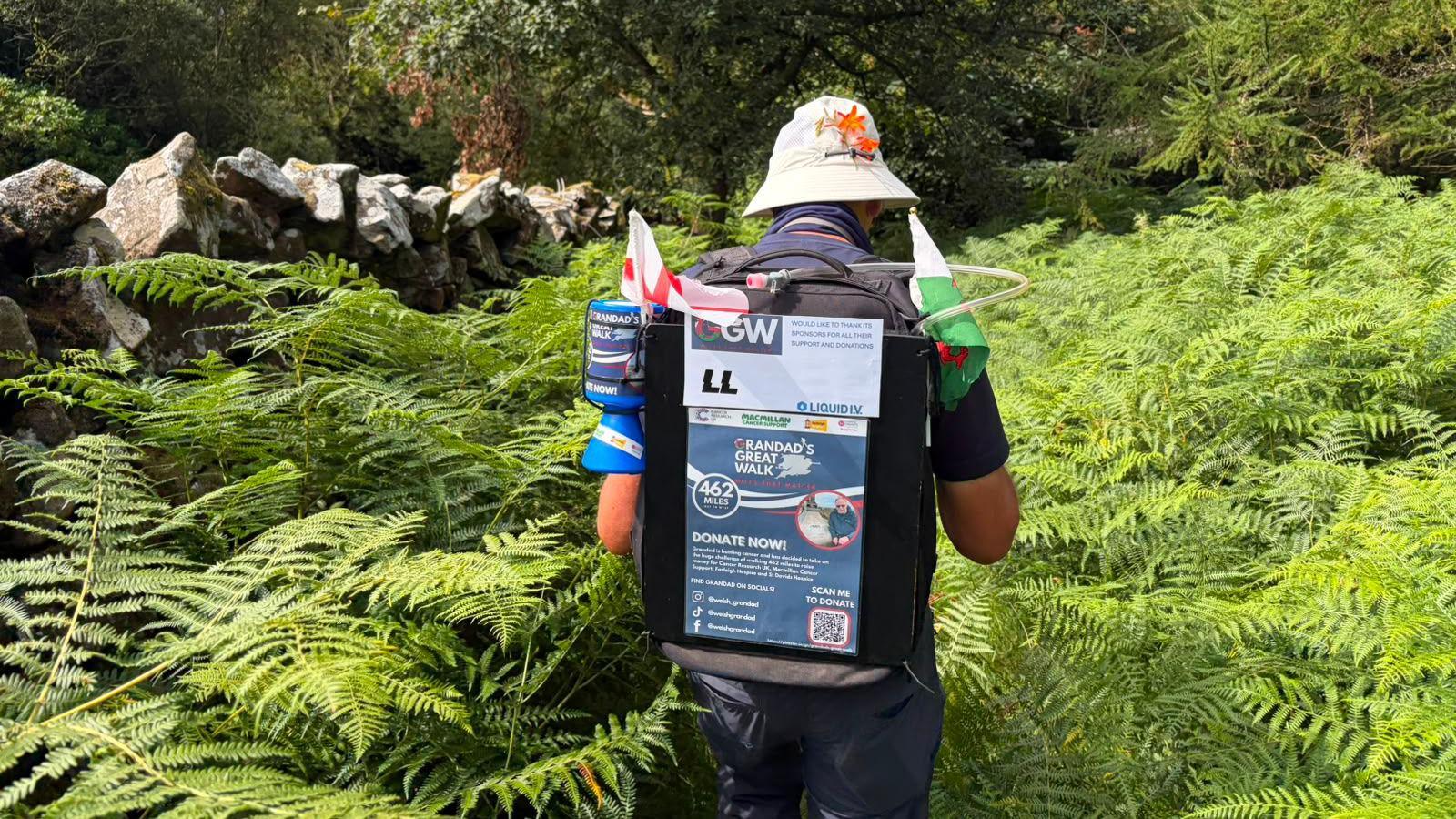 John walks away from the camera through an overgrown footpath full of ferns. He is wearing a backpack that is decorated with a Welsh flag and English flag, as well as the information on how to donate to his fundraising walk.
