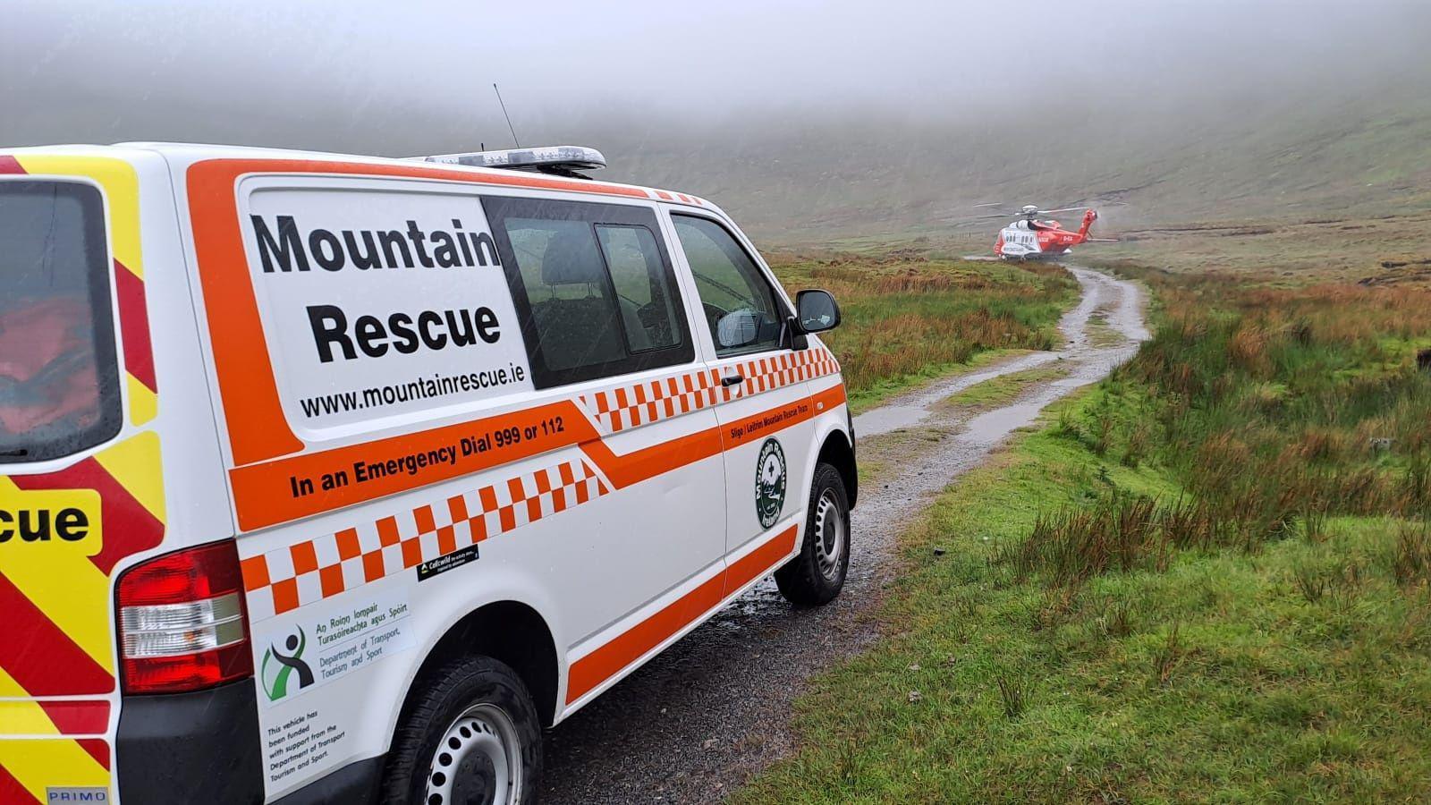 SLMRT response vehicle with ICG Rescue 118 helicopter on standby near the base of the mountain. The vehicle is white with orange pattern. The helicopter is white and orange. There is a path and grass in the foreground with mountains in the background. It is cloudy.