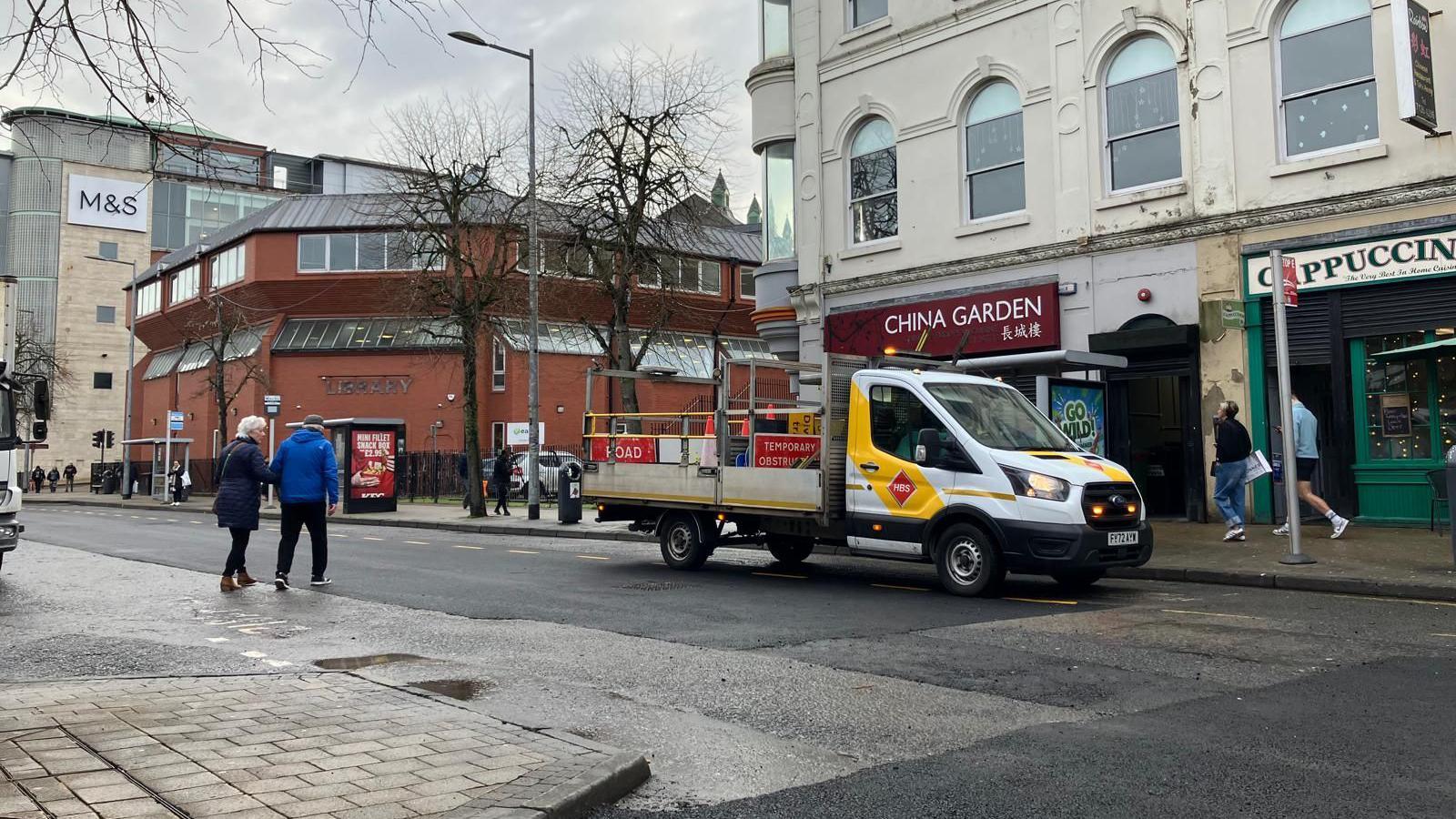 A construction truck sits parked on Foyle street in Derry, outside a takeaway with the red brick library and the marks and spencer store seen in the background. A man and a woman can be seen crossing the street