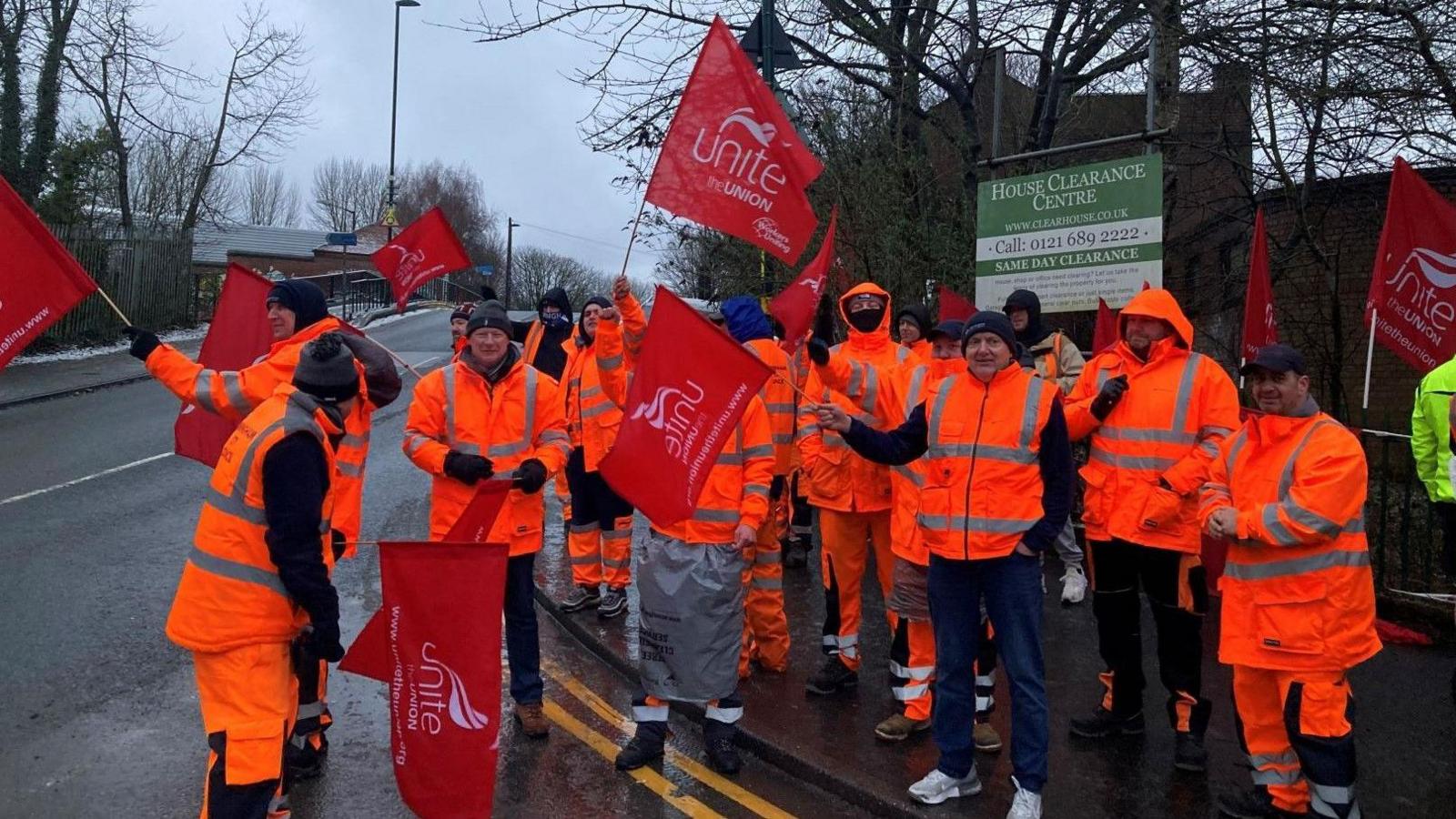 A group of men wearing high-vis orange jackets and trousers stand on a picket line waving red Unite the union flags, next to a road near the canal.