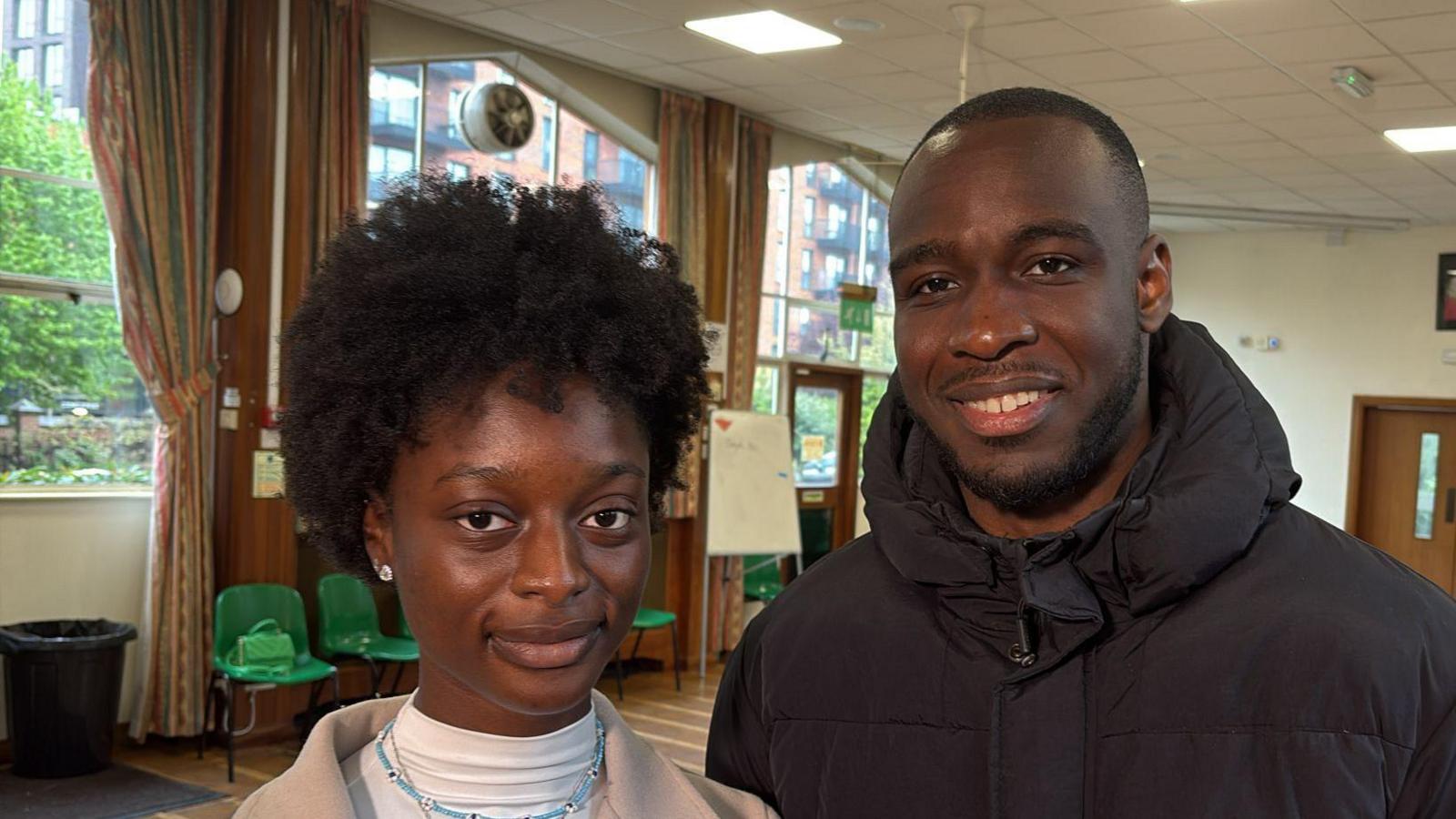 A woman and a man stand in a hall, smiling at the camera.