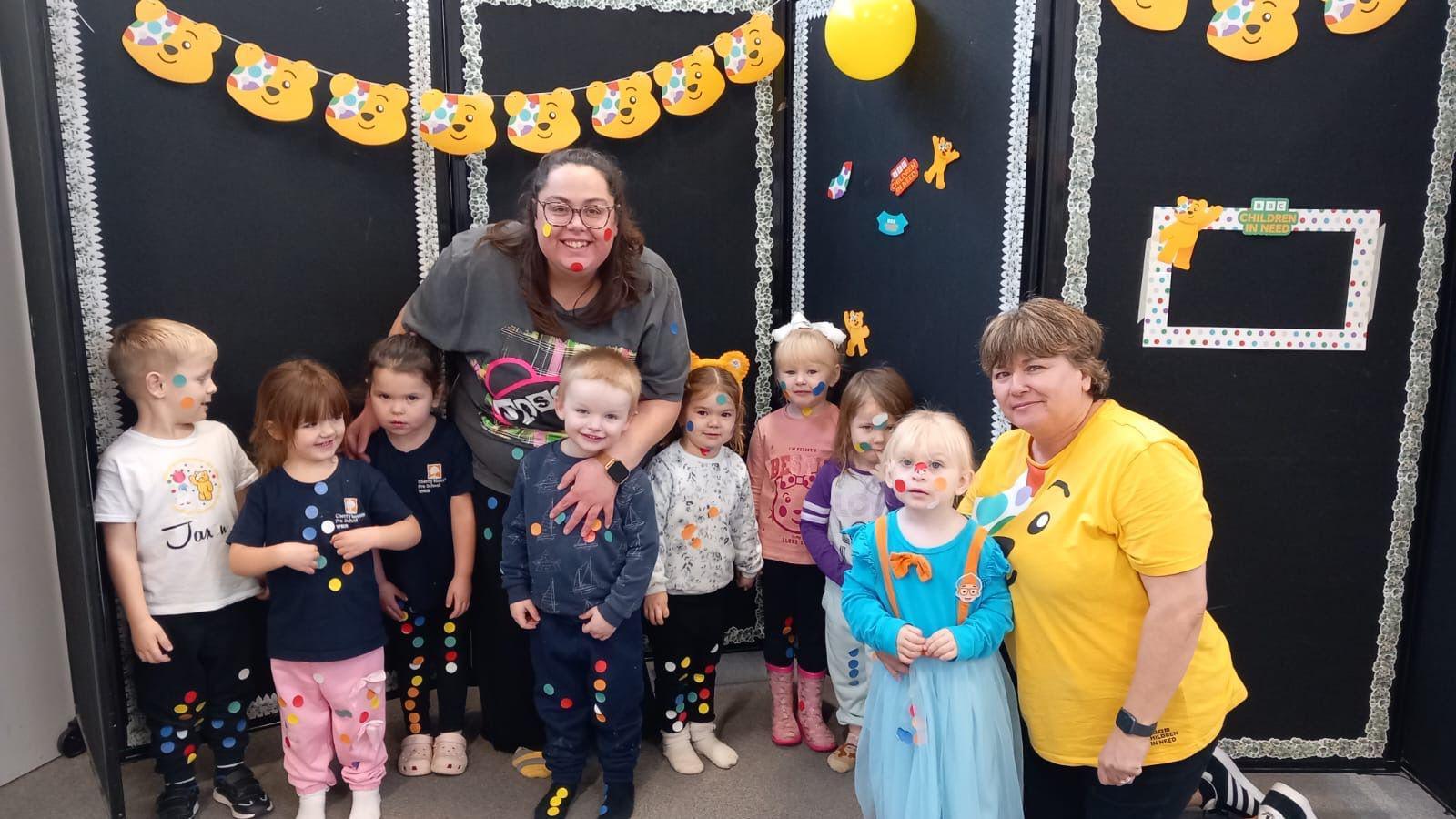 A group of eight children, all under the age of four, stand in a line with two pre-school workers. They all have stickers on their faces and clothes and are standing in front of a banner with Pudsey bear faces on it.
