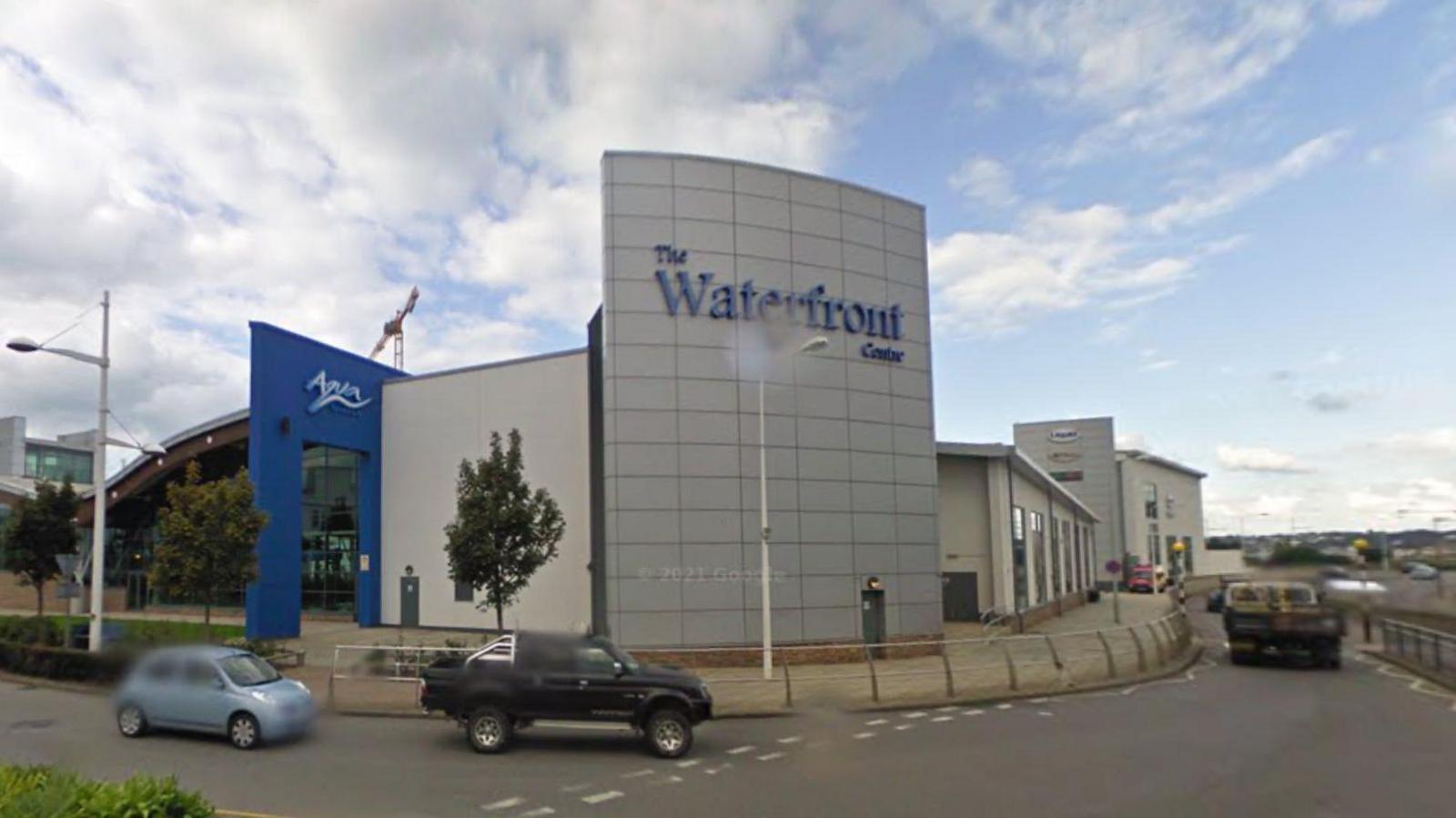 A picture of the front of the leisure centre. It is a large modern building with a main road in front of it. Cars can be seen driving past in the image