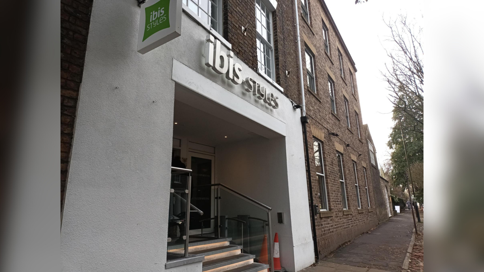 An exterior shot of Ibis Style Hotel in York, and red-brick building with a white plastered facade around the entrance, and the green Ibis style logo sign above. In the entrance, a short flight of stairs can be seen, alongside an accessible lift which has two orange cones placed in front of it.