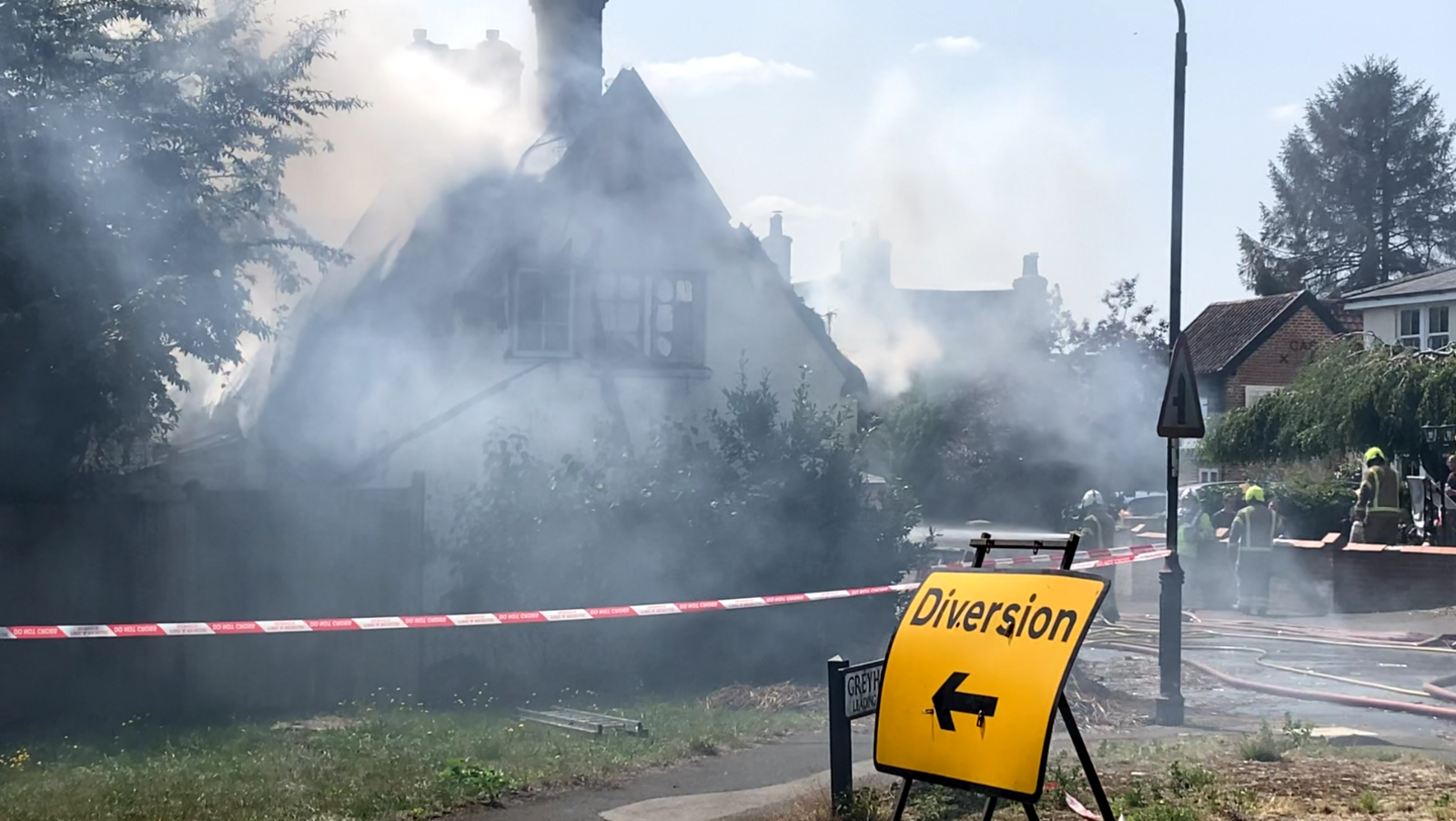 Lots of smoke is coming out of a badly damaged thatched cottage. A yellow Diversion sign is also in the picture, as are at least four firefighters.