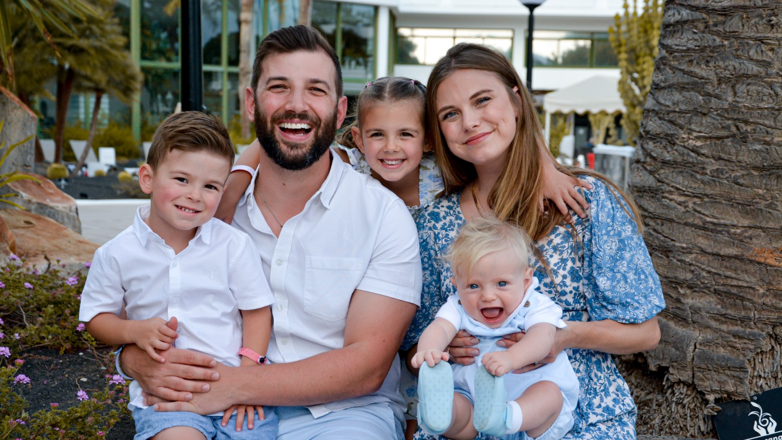 A man with dark hair and beard in a white shirt sat next to a woman in a blue and white floral dress. The man is holding a young boy on his lap also in a white shirt. The woman is holding a baby in a white shirt on her lap. Between the adults is a young girl's head. They are sat next to a tree trunk