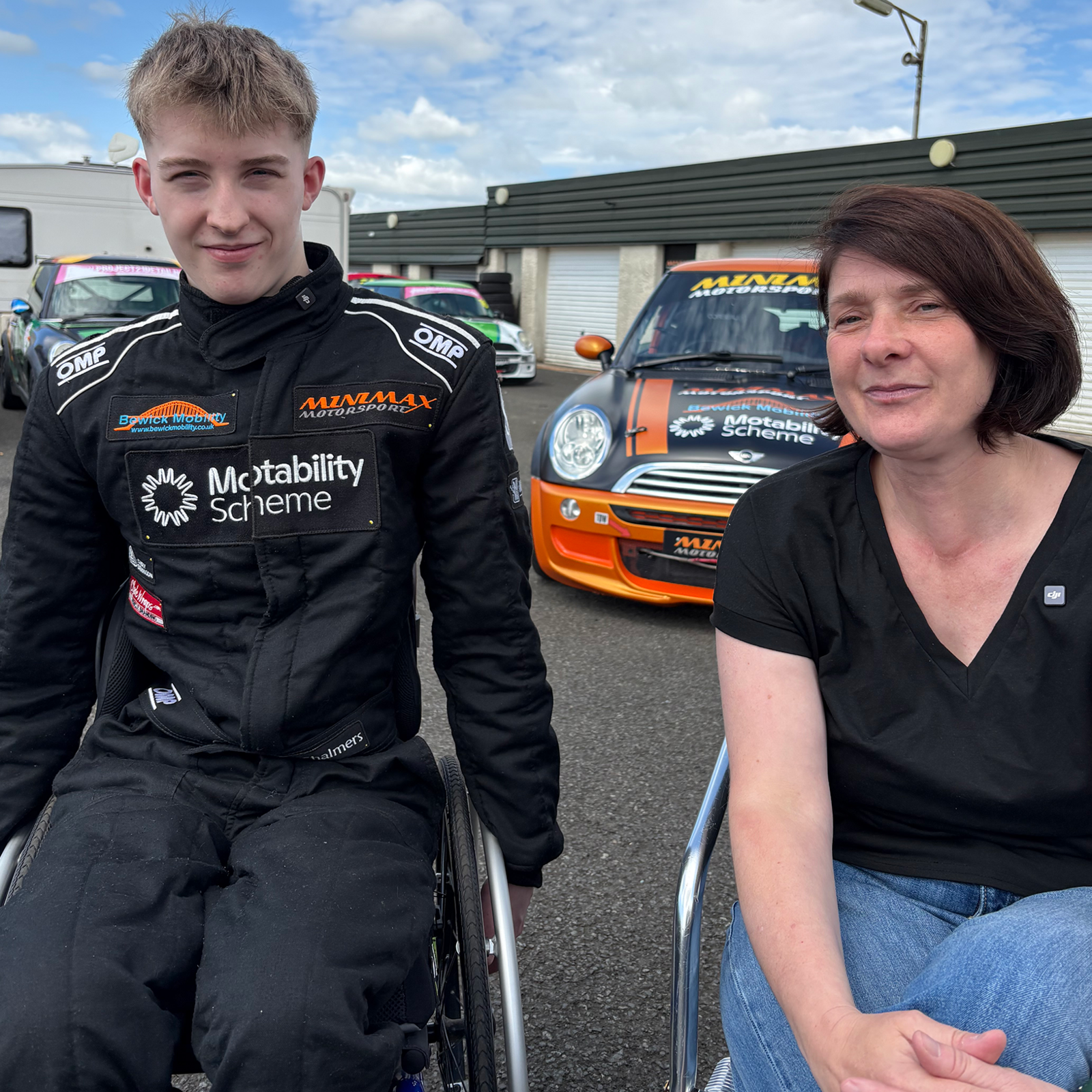 Ally Chalmers, a 17-year-old boy with short, blond hair next to his mum. He is in a wheelchair and is looking at the camera and half smiling. He is wearing a black race suit with the names of sponsors on it. His mum has short, brown hair in a bob style. She is wearing a black v-neck T-shirt and blue jeans. There are racing cars behind them.