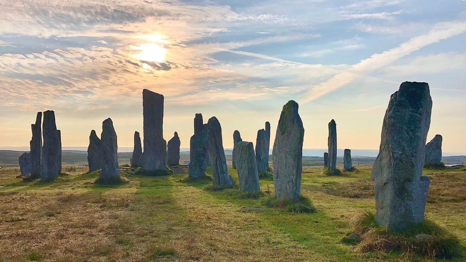 A general view of the Callanish Stones. There are a number of grey standing stones of varying sizes. All of the stones are grey, but those in the background of the image look darker due to the shadows of the sun. The sun is above the tallest stone and is partially obscured by cloud. The sky is blue. To the right of the image, two clouds have crossed over in the sky in a Saltire pattern.