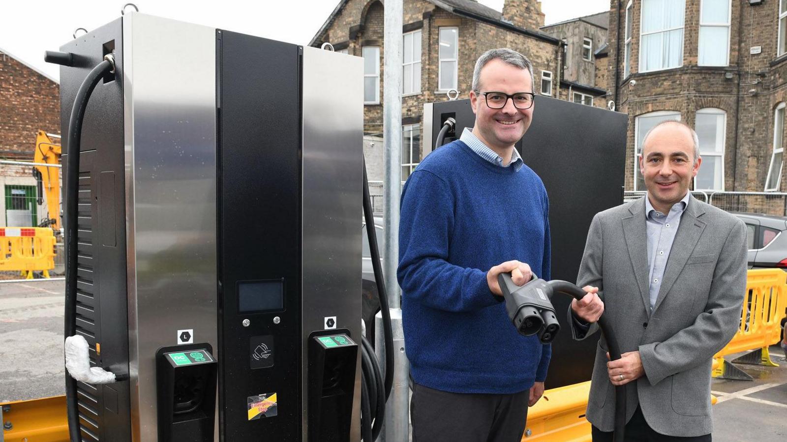 Two men stand together holding a bus charging cable next to a bus charging point. The man on the left is taller, wearing a blue jumper and glasses. The man on the right wears a grey suit jacket and shirt. They are both smiling.