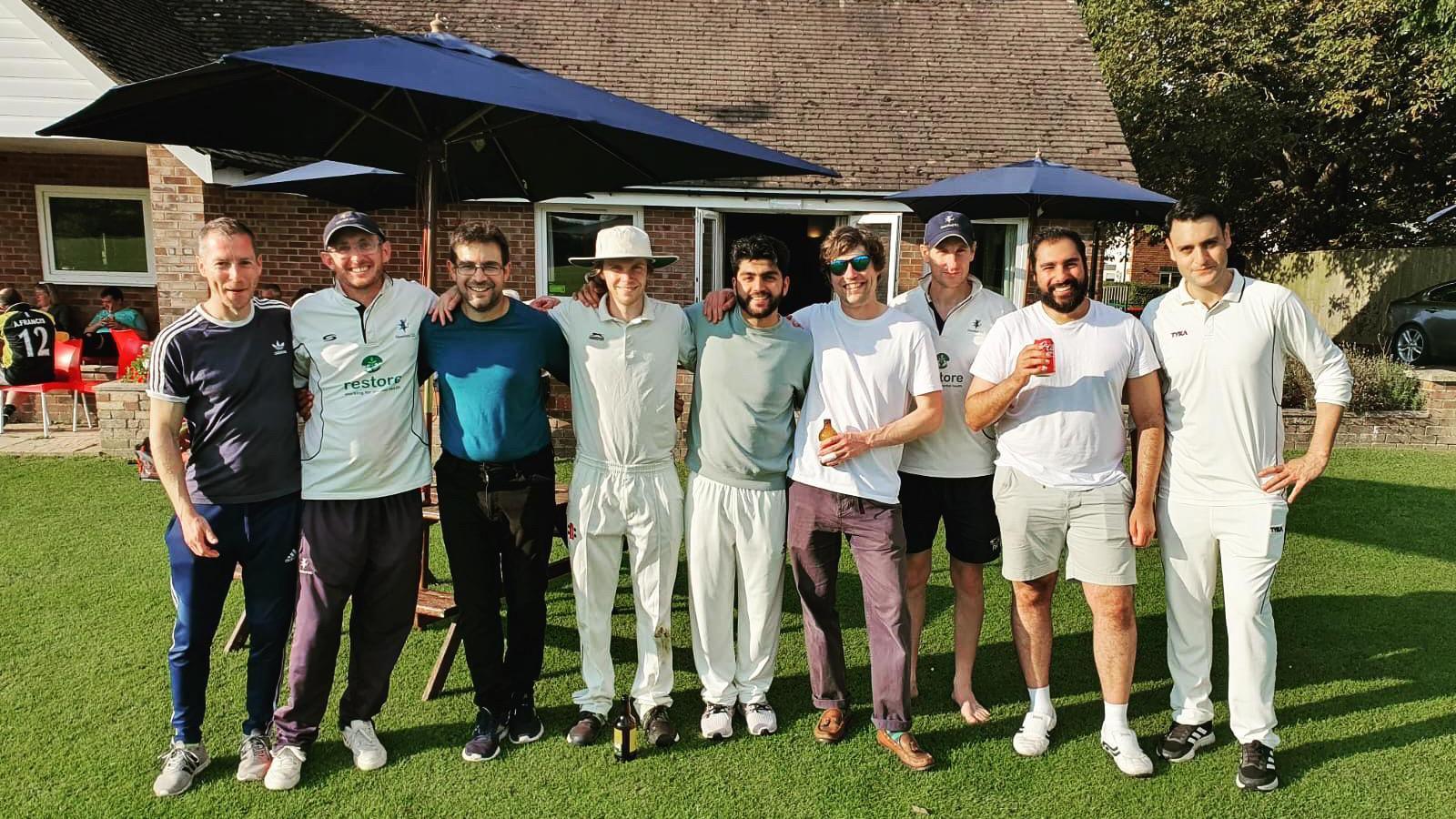 Nine men cricket players in casual clothes smiling for the camera in front of a pavilion. Some are holding drinks. It's a sunny say.