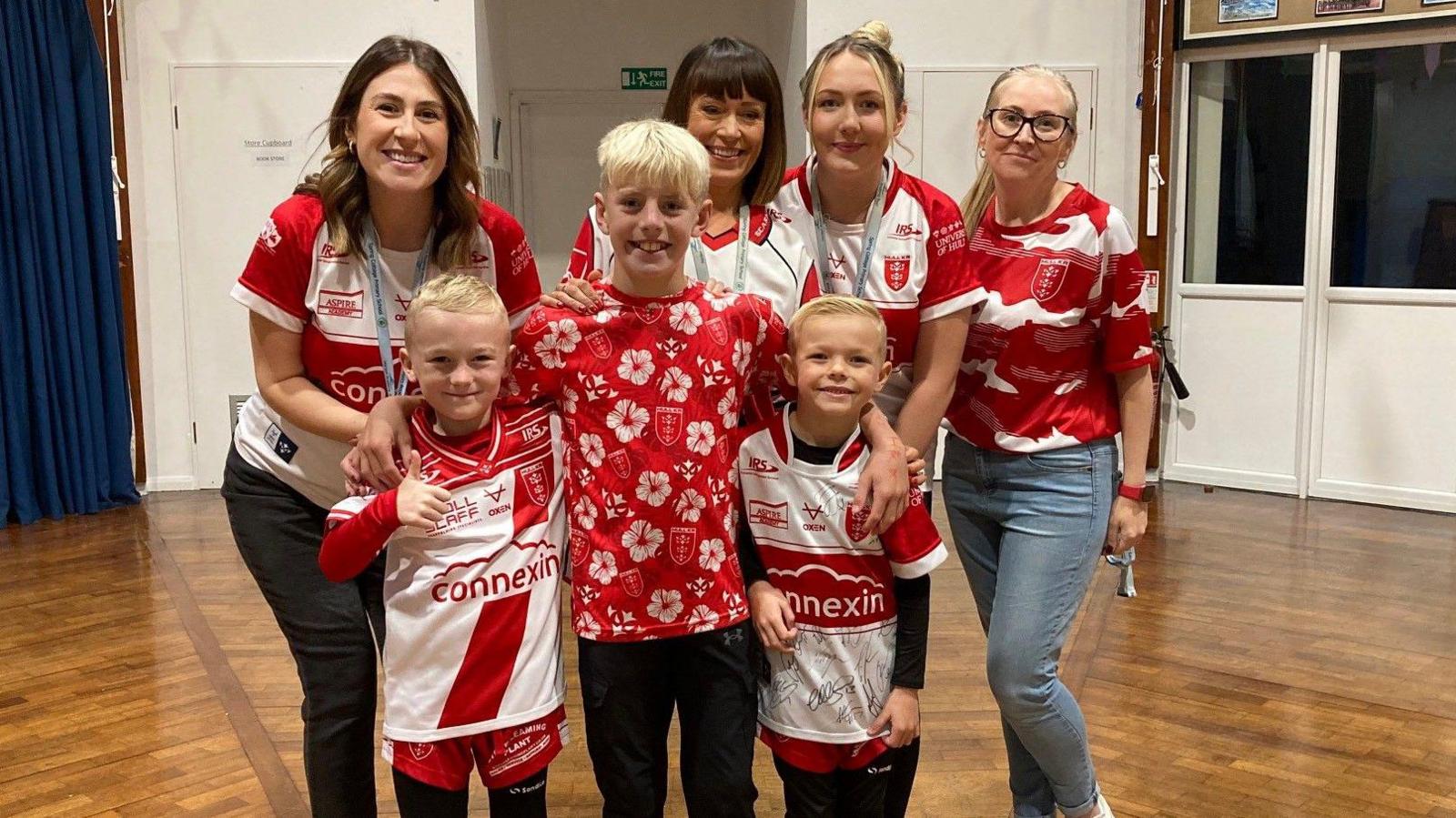 Three children, wearing red and white shirts, stand with four members of staff who are also wearing Hull Kingston Rovers memorabilia. They are in a school hall surrounded by examples of the children's work.