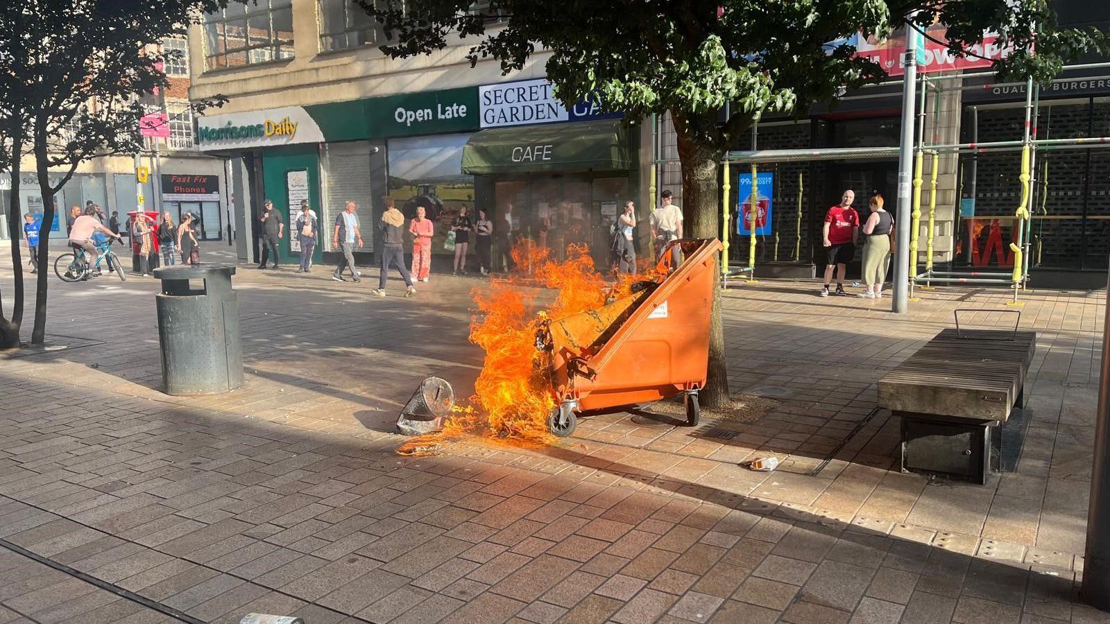 An orange wheelie bin on fire in the city centre. Morrisons shop is in the background along with scaffolding on a nearby building. About 15-20 people are gathered in the background