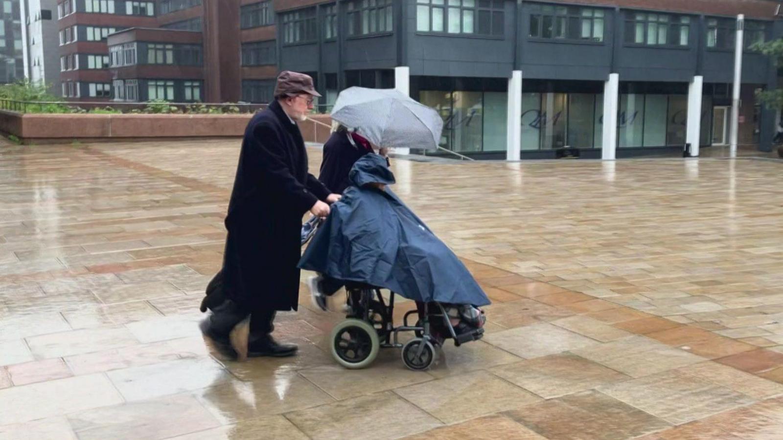 June Mills, whose face is covered by a dark blue waterproof poncho, is pushed away from Liverpool Crown Court in her wheelchair by her husband 