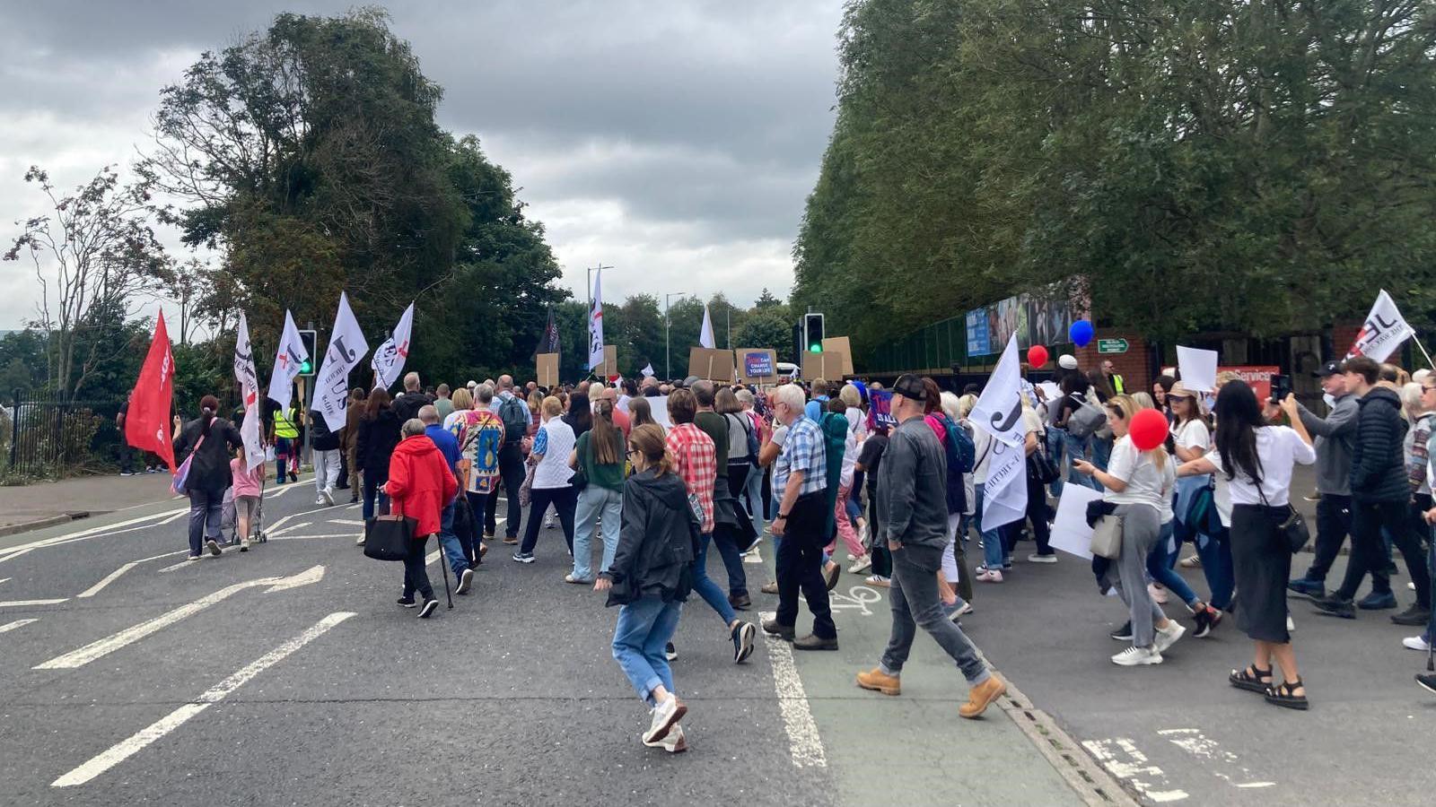 A group of people waving flags leave the side of Ormeau Park, on the Ormeau Embankment.