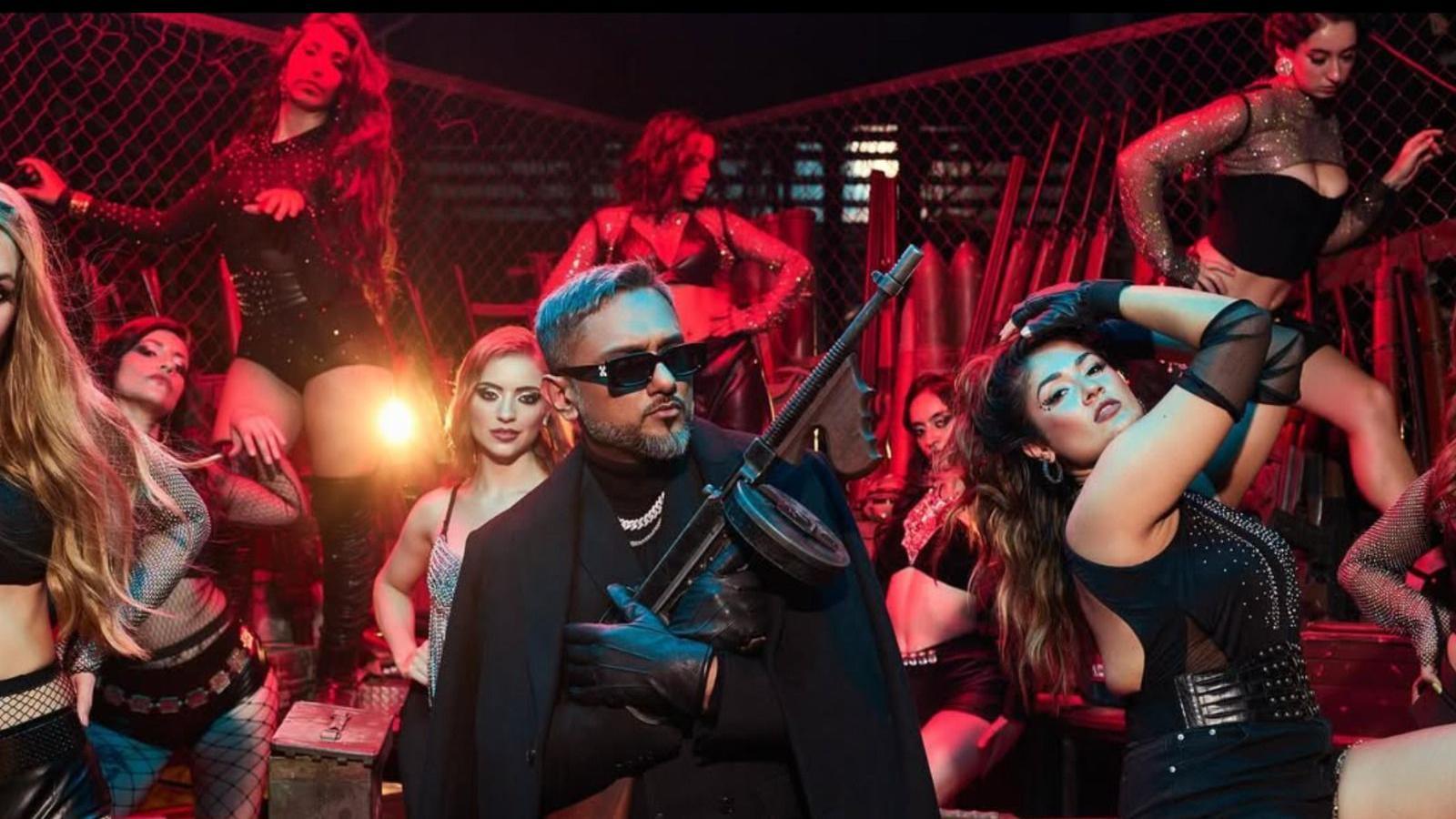 A picture of Yo Yo Honey Singh wearing a black jacket over a black suit and a silver chain with black leather gloves. He is holding a gun and has background female crew wearing black dresses 