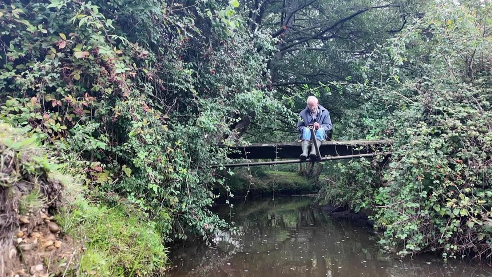 Mike James seated on the small concrete bridge over a stream with 