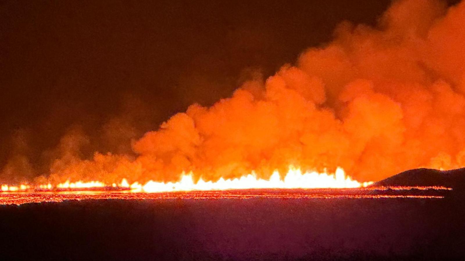 A line of flamed on the horizon, with orange smoke billowing from it into the night sky and a line of molten lava flowing in front