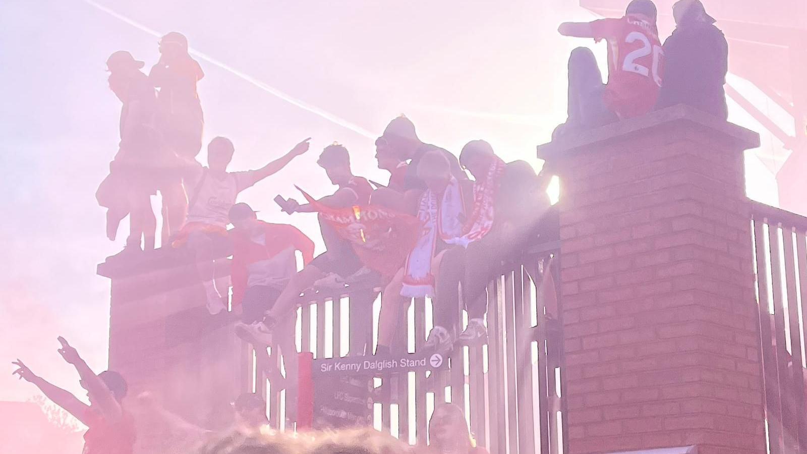 Fans adorned in Liverpool scarfs and carrying flags sit atop a fence outside Anfield stadium.