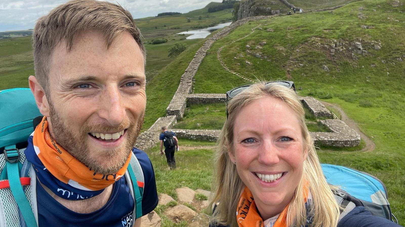 Luke Hames-Brown has a mousey brown beard and short hair and Kate has blonde, shoulder length hair. They're both carrying rucksacks and wearing MND Association branded snoods