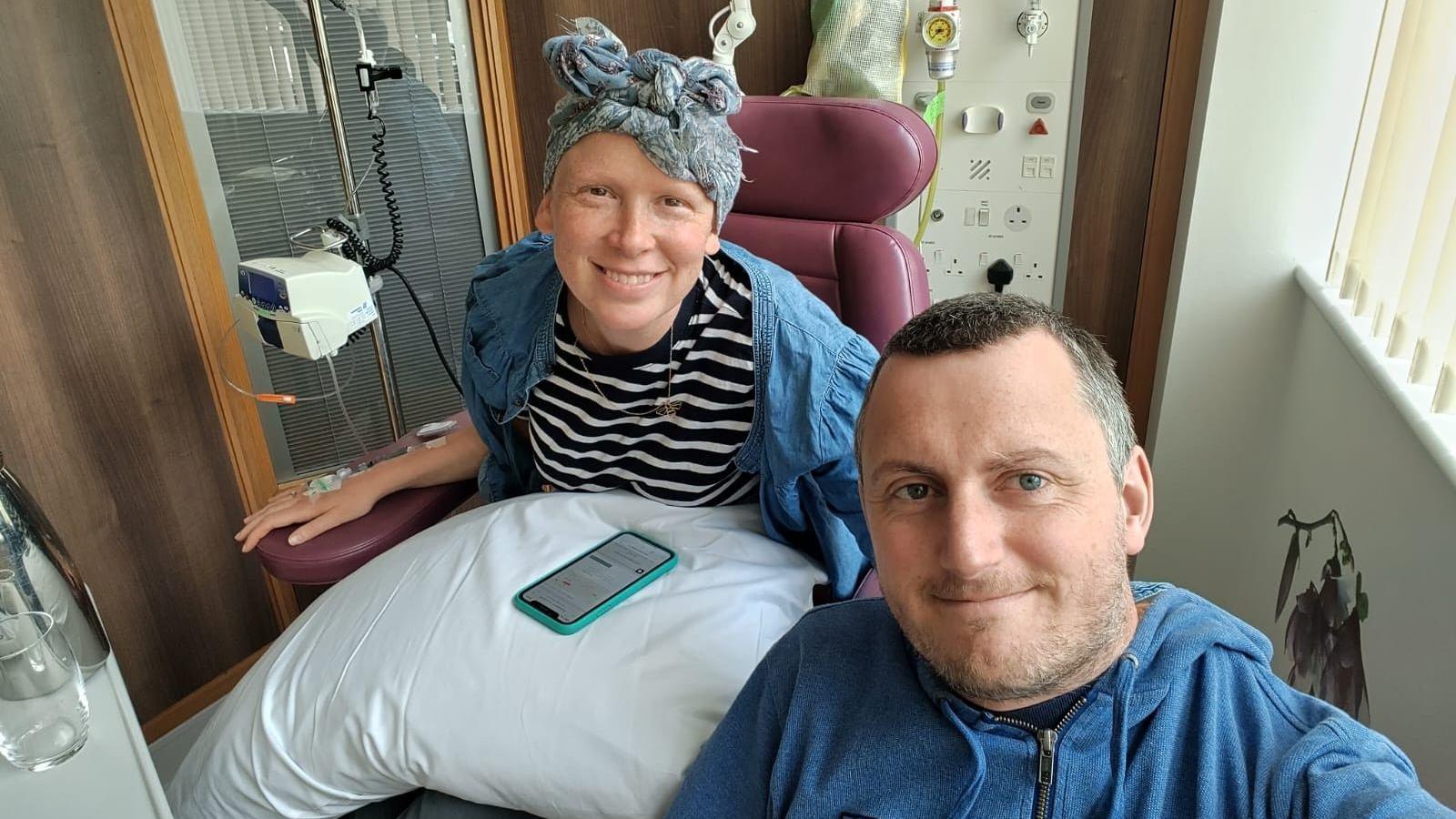 Phillippa Hentsch and her husband Matt. Phillippa is sitting in a chair with a cannula in her hand. She has fabric tied on her head and a phone on her lap.