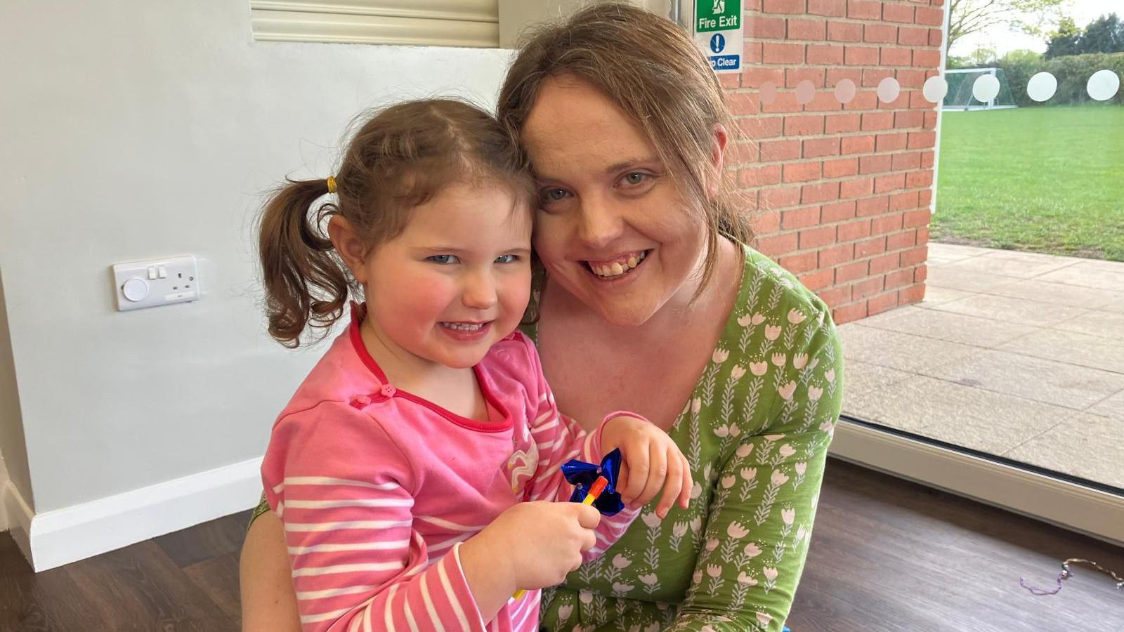Rachel and her daughter Mia sit on foam play mats indoors. Mia is wearing a pink top and sits on Rachel’s lap holding a small toy. Behind them are a glass door, brick wall and outdoor grass.