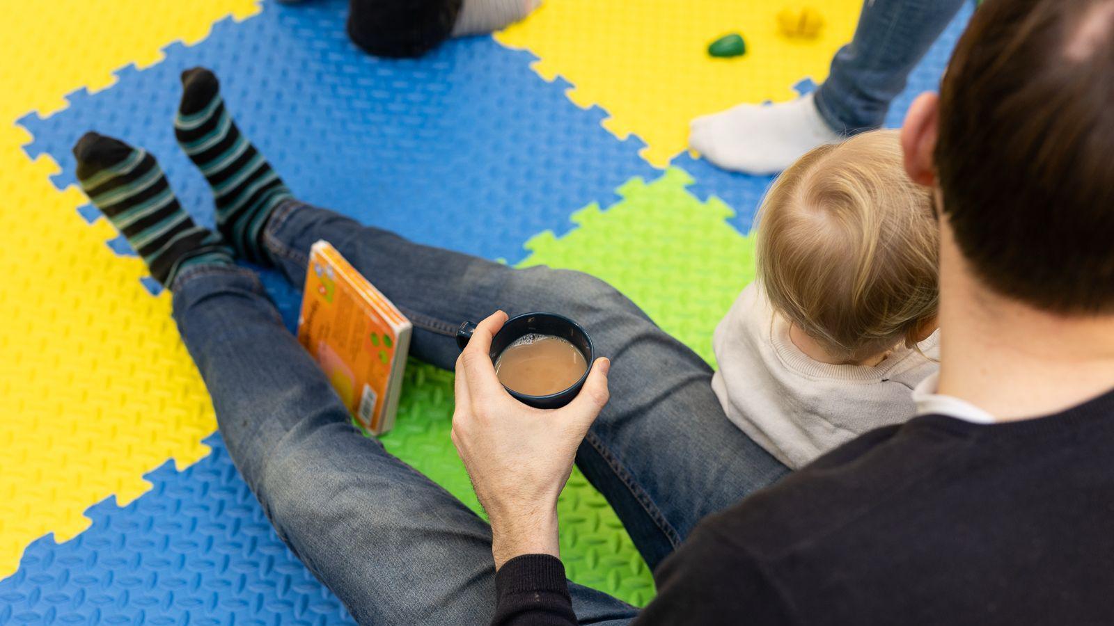 A father and his child sit on a colourful foam panelled floor, the father is holding a cup of tea and has a children's book resting between his legs.