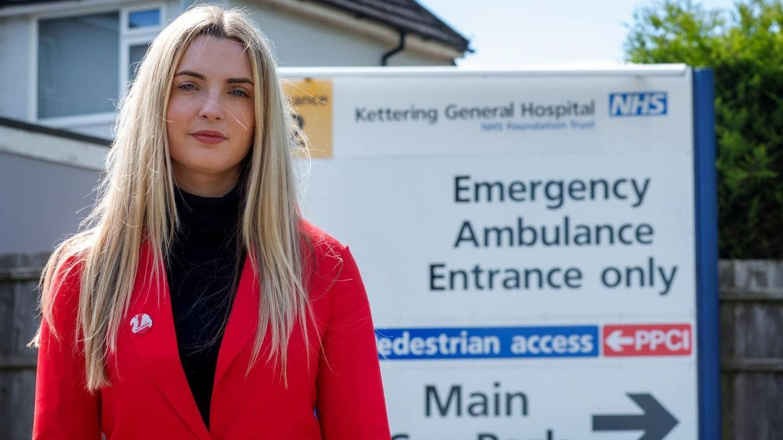 Rosie Wrighting in a black roll neck and a red blazer stands by a sign for Kettering General Hospital. 