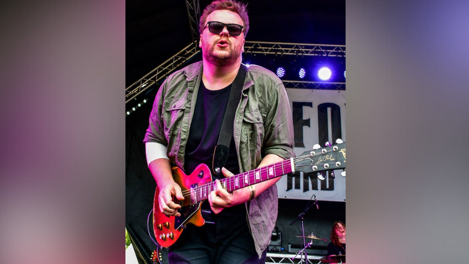 A picture of Matt Long playing guitar on stage at Alfold Festival in Surrey. Matt is wearing sunglasses, a khaki shirt, black t-shirt and jeans. 