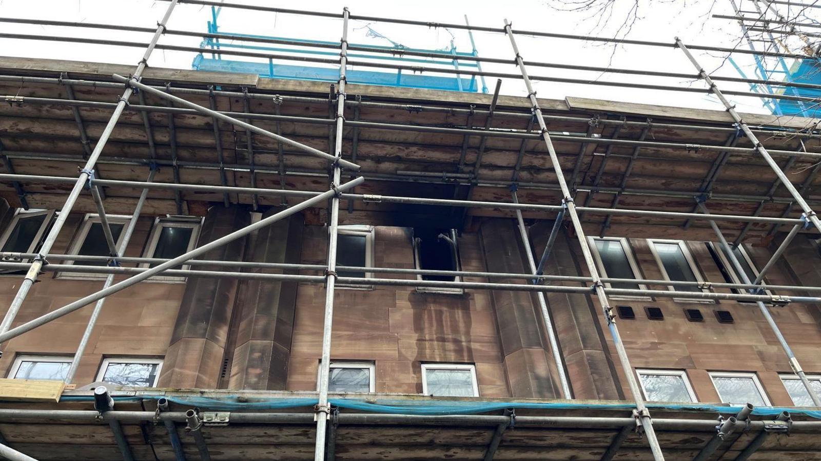 A close-up image of a brown building with narrow rectangular windows. One of the windows has no glass in it, and has black stains on the walls around it. There is scaffolding on the building.