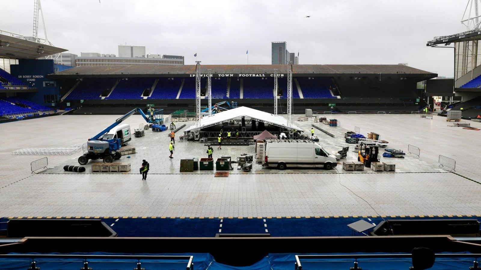 A large metal structure with a white roof, which all looks like a big tent, is in place in the middle of the pitch. It is surrounded by the football stadium's stands. Workers in hi-vis jackets are working around it and various vehicles are parked nearby. The pitch is covered in white protective boards.