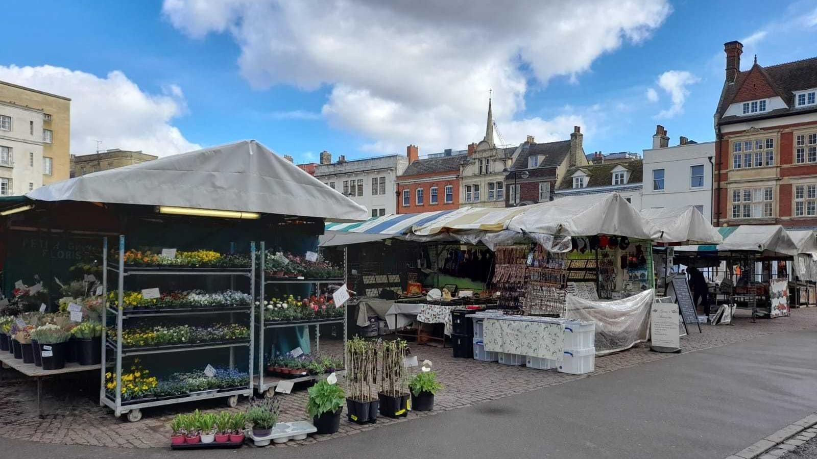 The photo is of the Cambridge market. The sky is blue with some clouds and the camera is mainly focused on a plant stall with a grey tarp roof. There are buildings of various colours in the background and rows of stalls behind the plant stall which have white, blue, yellow or green striped tarp covers atop. 
The stalls are on cobbled flooring. Around the market is a smoother concrete path.