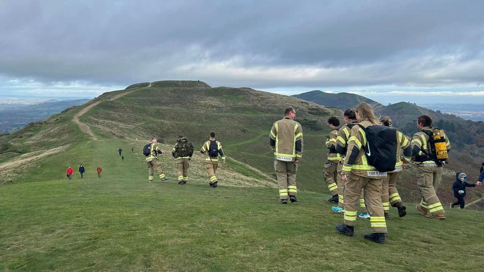 A group of people in firefighting uniform photographed from behind on a hill ridge with a series of peaks in front of them into the distance.