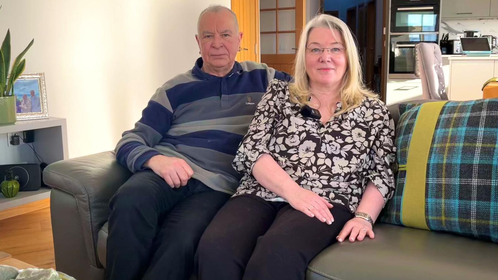 Asa and Kathleen are sitting on a sofa in the living room of their home. It is a bright and airy room.