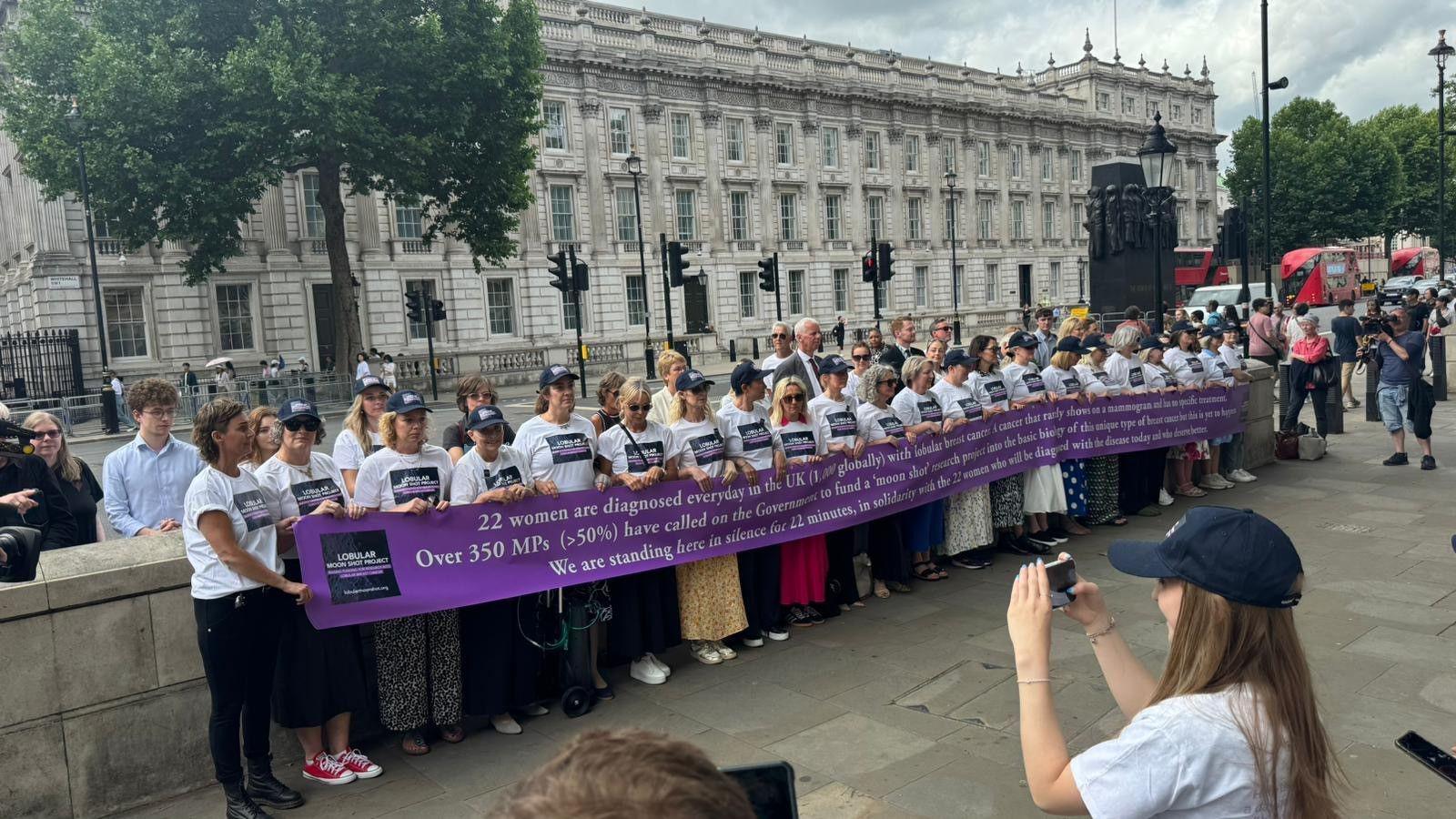 A group of women, all wearing white campaign t-shirts, stand in front of a set of white Portland stone government buildings in Westminster, all holding a long purple and white campaign banner.